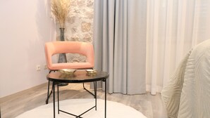 A modern room with a pink chair, a small table, a vase with dried plants, and a view of a stone wall through a window with grey curtains.