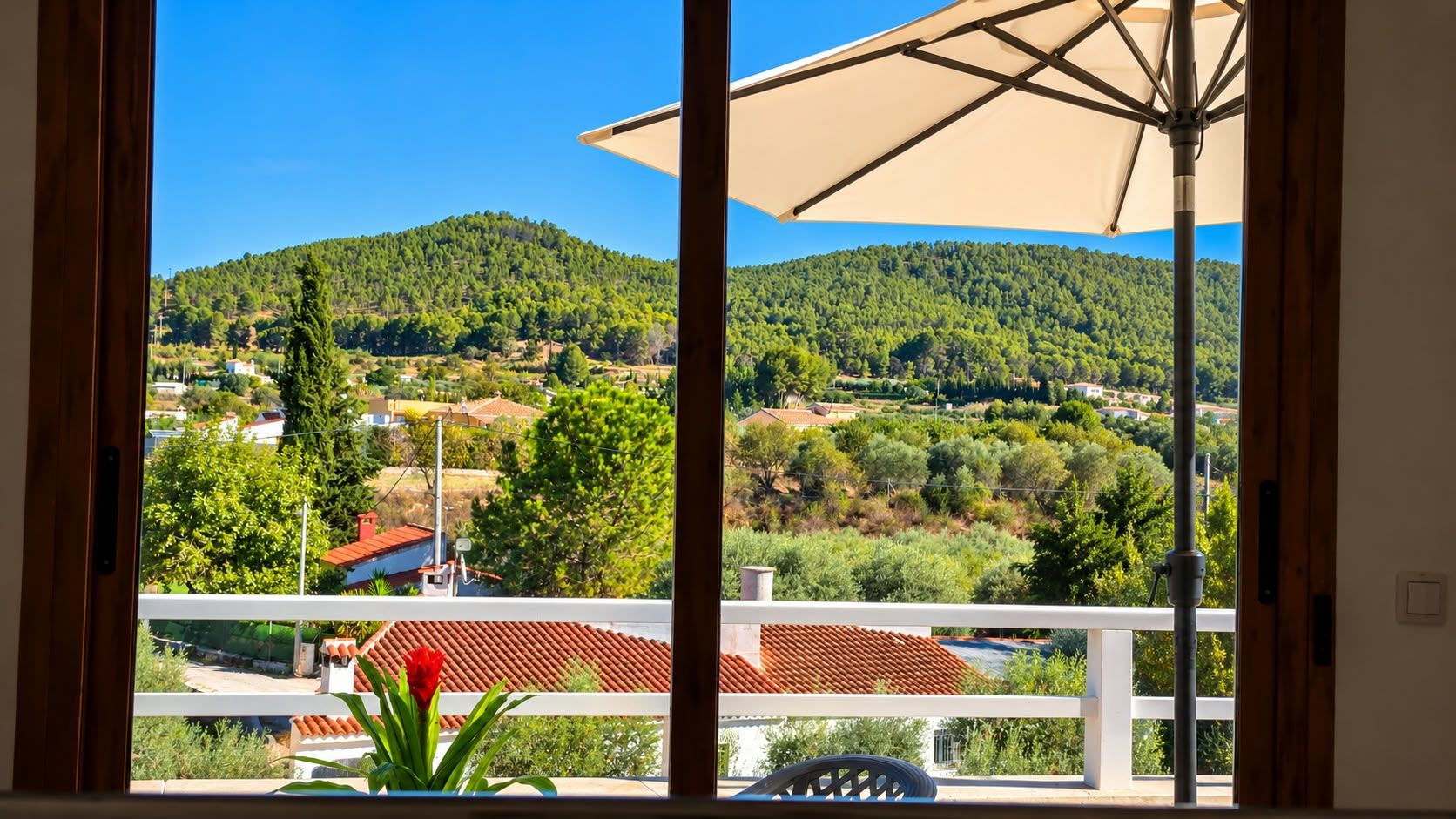 Un balcón con vistas a un paisaje montañoso, una mesa con una flor roja y una barandilla blanca.