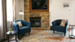 A living room with a brick fireplace, two armchairs, a floor lamp, and a colorful rug.