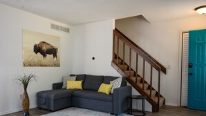 A living room with a gray sectional sofa, a colorful rug, a wall-mounted picture of a bison, and a wooden staircase.