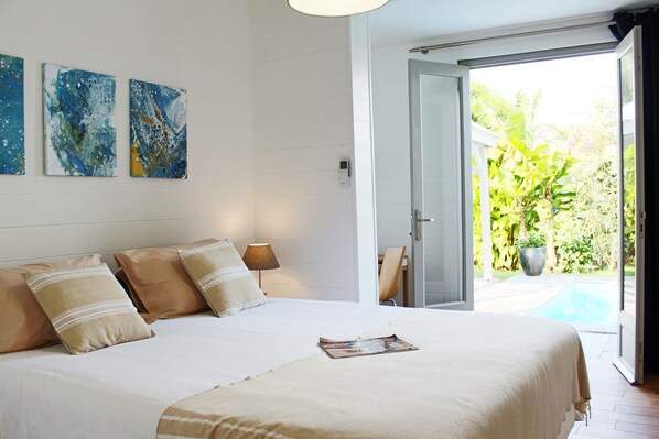 A bedroom with a bed, pillows, a bedside table, a lamp, and a view of a garden and pool through an open door. - Villa le Sucrier (Les Trois Ilets)