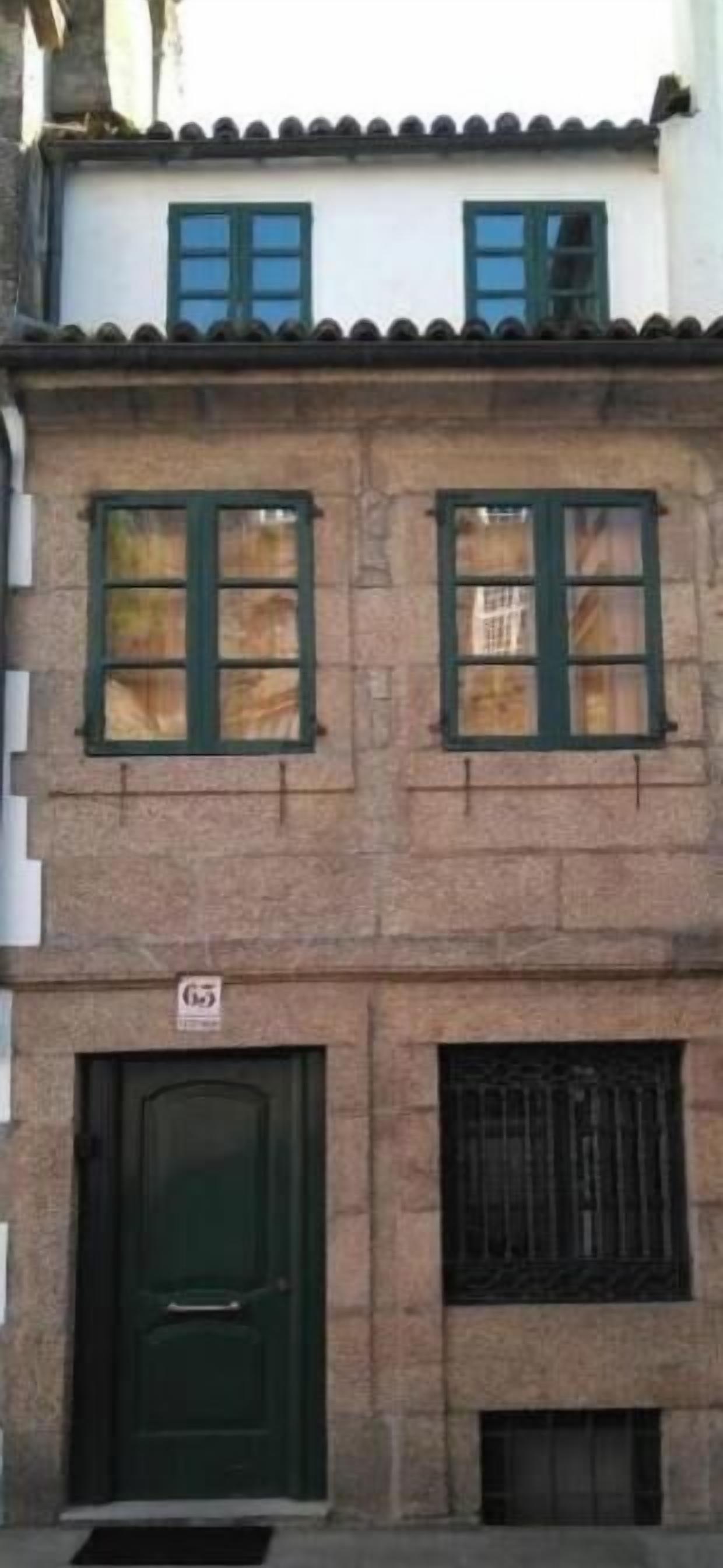 Un edificio de dos pisos con una puerta verde, dos ventanas con contraventanas verdes y un balcón pequeño.