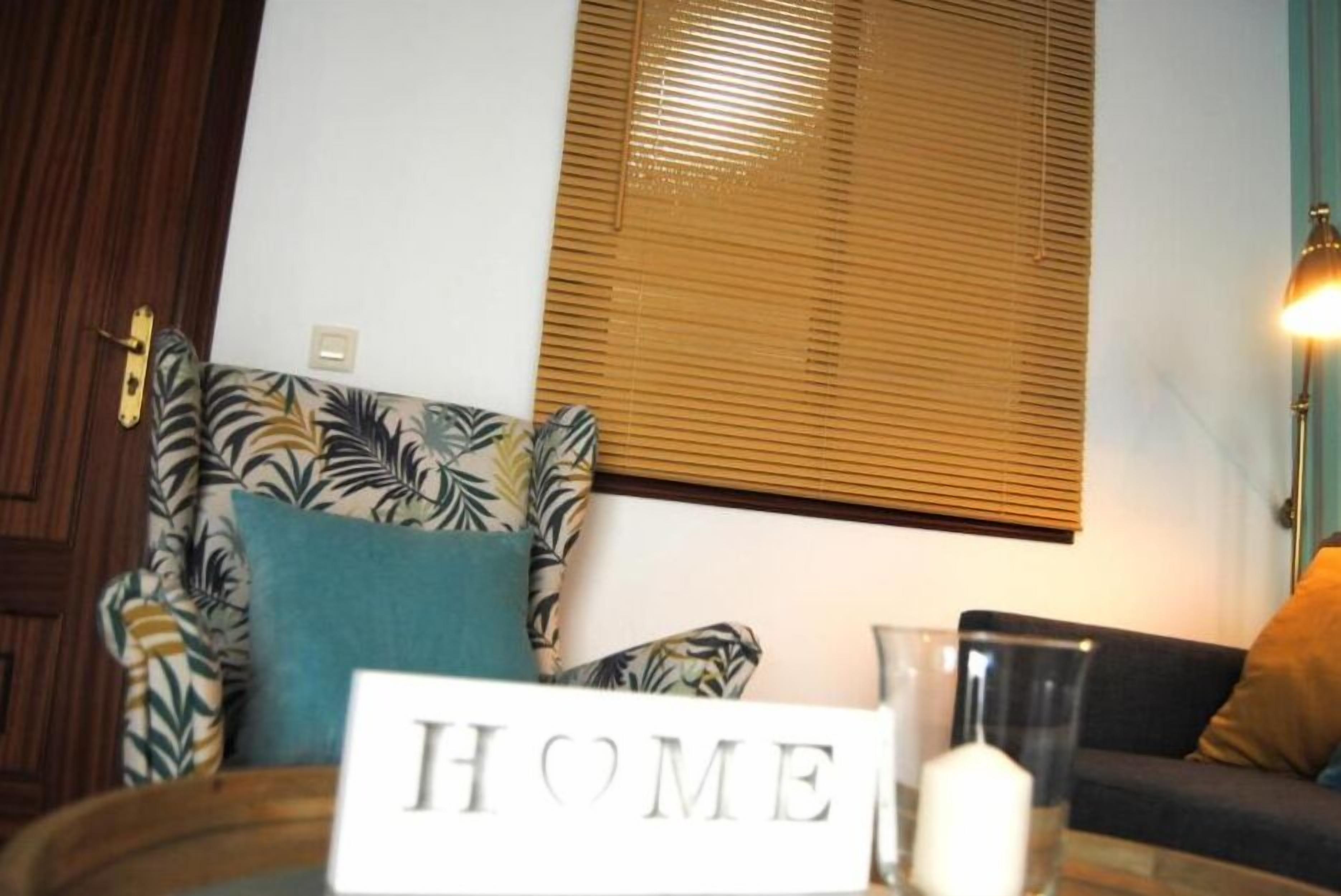 A living room with a sofa, a coffee table with a 'HOME' sign, a window with blinds, and a lamp.
