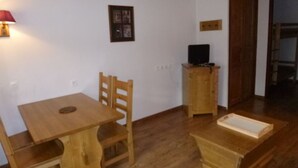 Um quarto pequeno e simples, com uma mesa e cadeira de madeira, um pequeno armário com uma televisão e um banco de madeira.