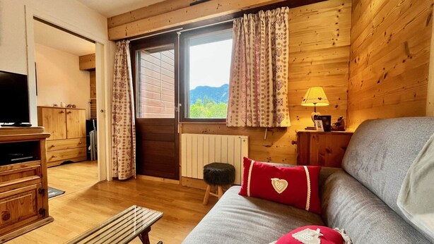 A cozy room with a wooden interior, a sofa with red cushions, a bench, a TV, and a window with curtains.