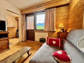 A cozy room with a wooden interior, a sofa with red cushions, a bench, a TV, and a window with curtains.