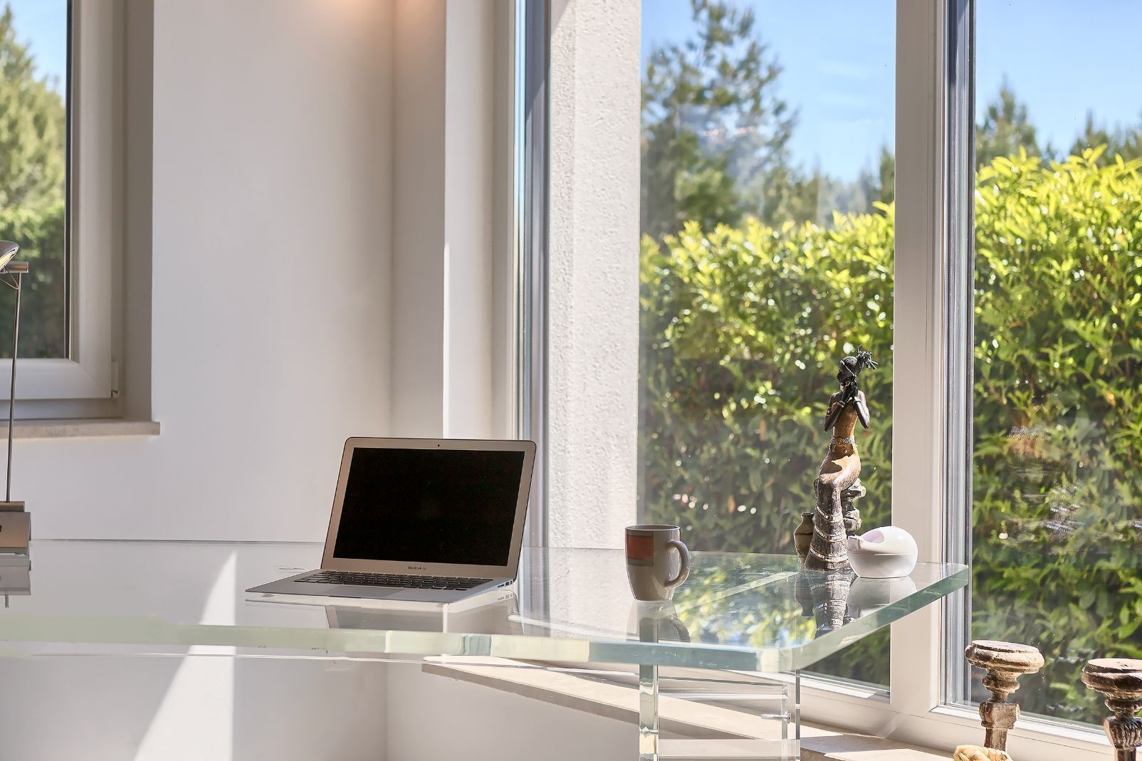 Una computadora portátil sobre una mesa de vidrio, cerca de una ventana con vista a la vegetación.