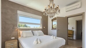 Ein modernes Schlafzimmer mit einem großen Bett, einer Chandelüster, einem Fenster und einer Tür zu einem anderen Raum.