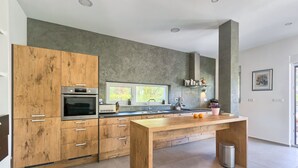 Eine moderne Küche mit Holzschränken, einem eingebauten Ofen, einem großen Holztisch und einem Fenster, das für natürliches Licht sorgt.