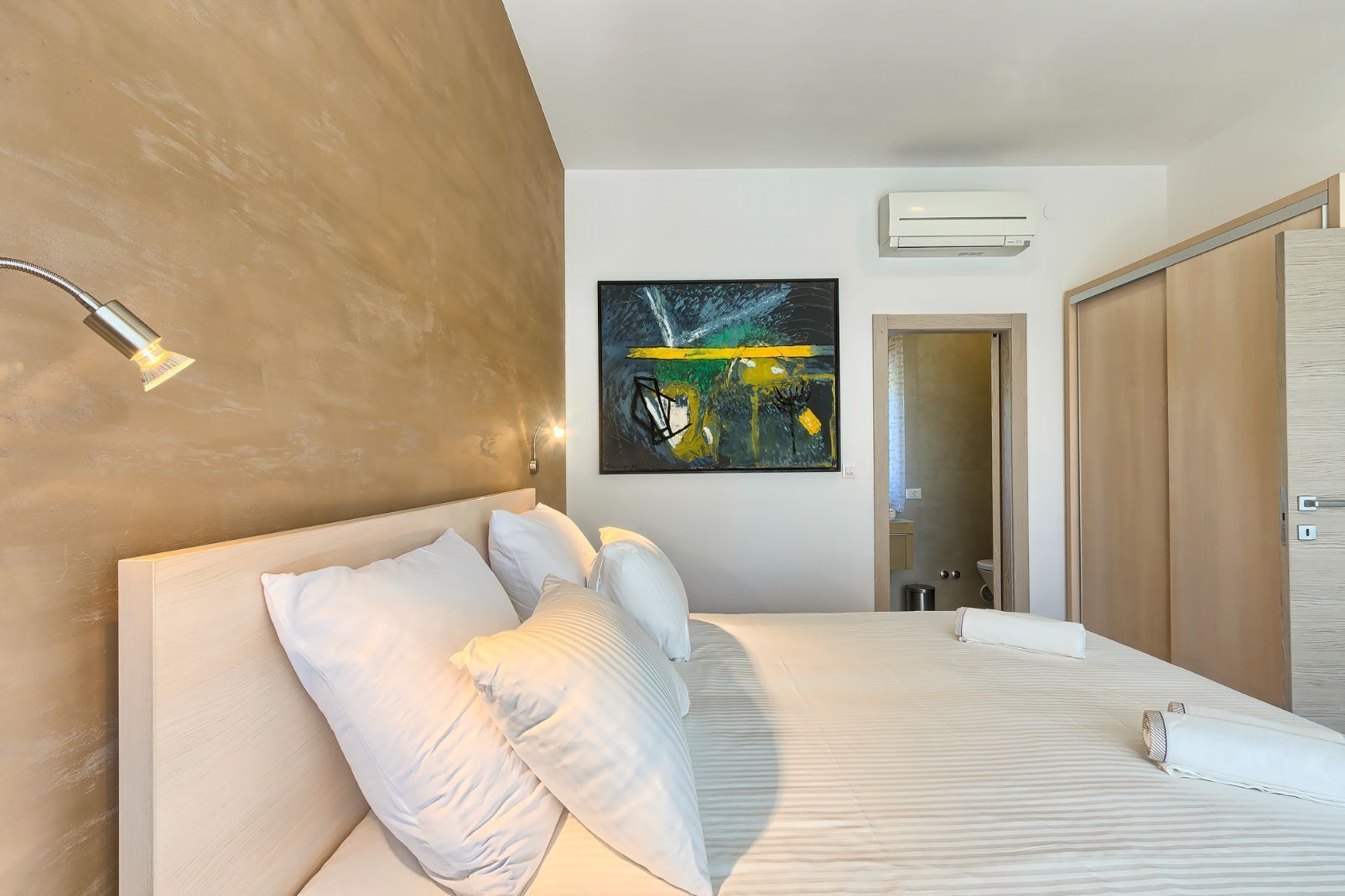 A modern hotel room with a large bed, a mounted artwork, an air conditioning unit, and a wooden wardrobe.