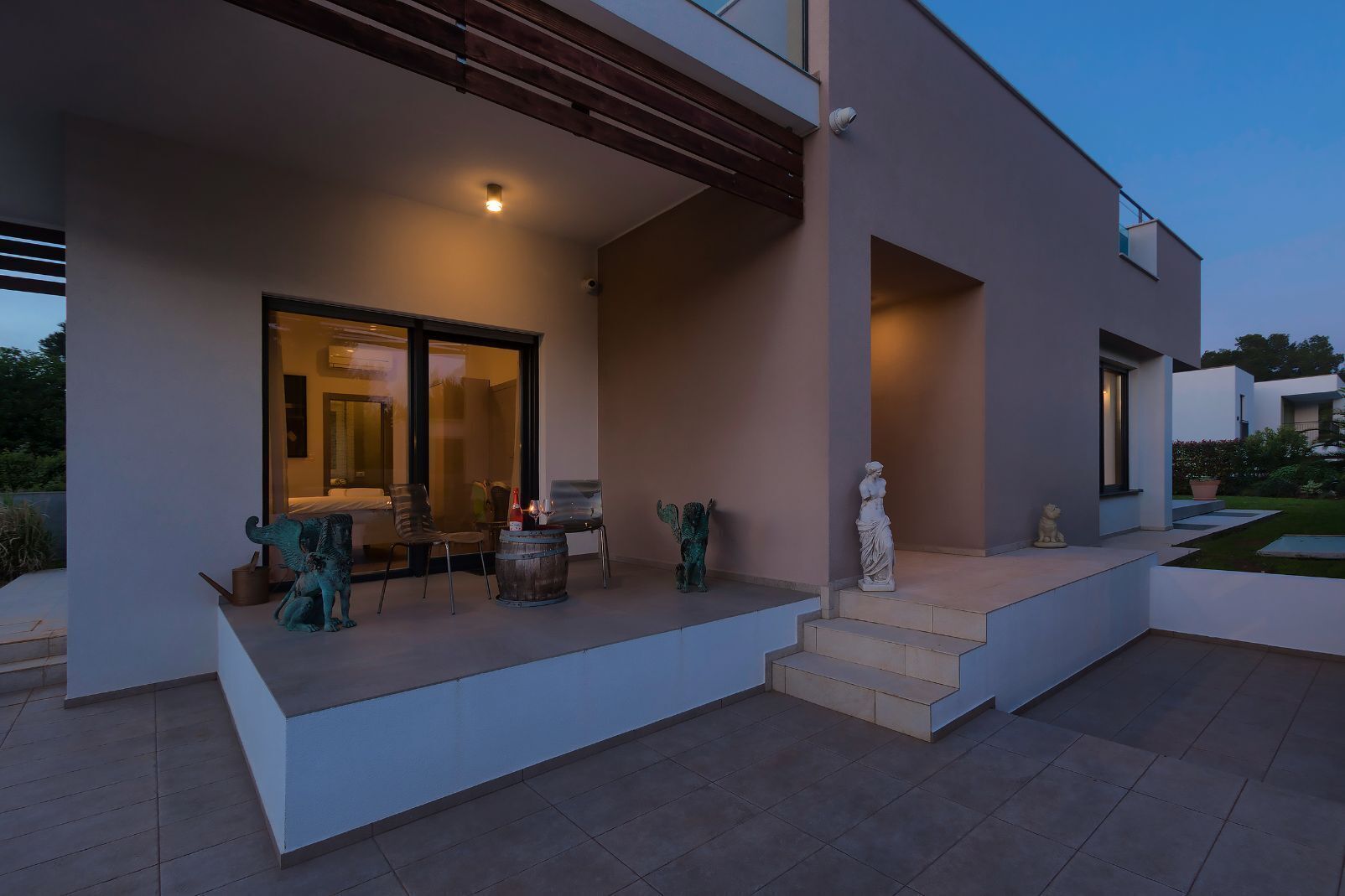 Une maison moderne dotée d’un patio couvert, d’un coin repas avec des chaises et une table, et d’une statue placée sur les marches.
