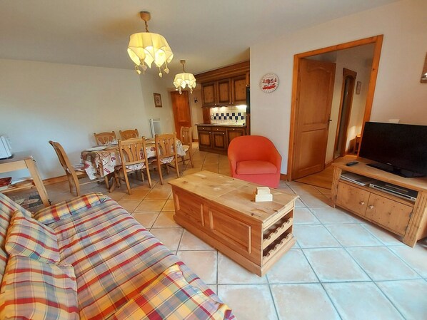 A cozy living room with a sofa, a coffee table, and a television. There is a dining area with chairs and a table, and a kitchen in the background. - T3 Apartment in Samoëns center with terrace and garage (Samoëns)