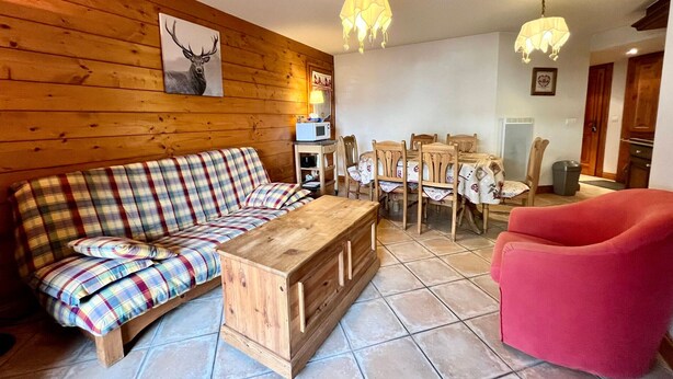 Un salon chaleureux avec un canapé à carreaux, une table basse en bois, un fauteuil rouge, une table à manger avec des chaises et un mur en bois orné d’un tableau représentant un cerf.
