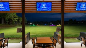 Ein Außenbereich mit Tischen und Stühlen, im Hintergrund ein Golfplatz und drei Bildschirme mit dem TopTracer Range Logo.