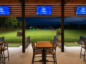An outdoor seating area with tables and chairs, a golf course in the background, and three screens displaying the TopTracer Range logo.