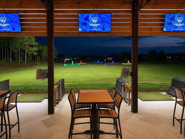 Un espace extérieur aménagé avec des tables et des chaises, un terrain de golf en arrière-plan et trois écrans affichant le logo de TopTracer Range.