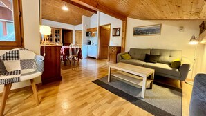 A cozy living room with a wooden ceiling, a sofa, a coffee table, and a dining area with wooden chairs and a table.