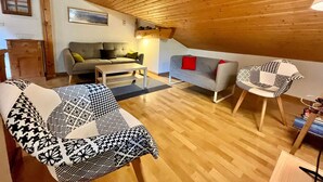 A cozy living room with a wooden ceiling, a grey sofa with red cushions, a patterned sofa, a wooden coffee table, and a patterned armchair.