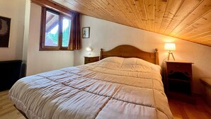 A bedroom with a wooden ceiling, a large bed, a nightstand with a lamp, and a window with curtains.