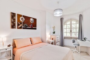 A bedroom with a bed, bedside tables, a desk, a chair, a lamp, and a window with curtains.