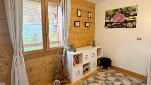 Een gezellige kamer met houten wanden, een raam met gordijnen, een klein wit kastje met boeken, een lijst met foto en een bloemschilderij aan de muur.