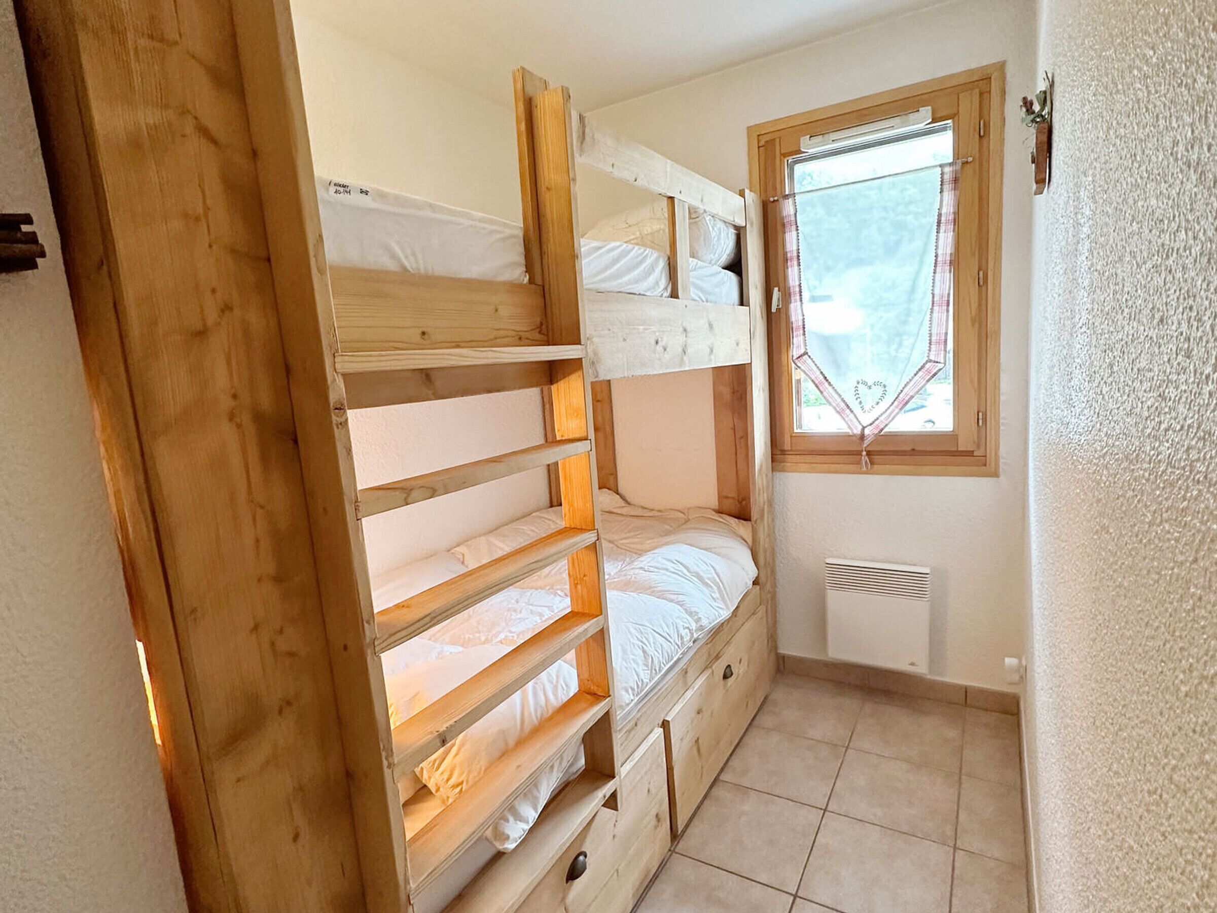 A compact room with a bunk bed, a window with a checkered curtain, and a radiator.