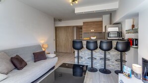 A modern living room with a sofa, a coffee table, and a kitchen area with a microwave and stools. - SUEDE  AP4207 by Riviera Holiday Homes (Niza)