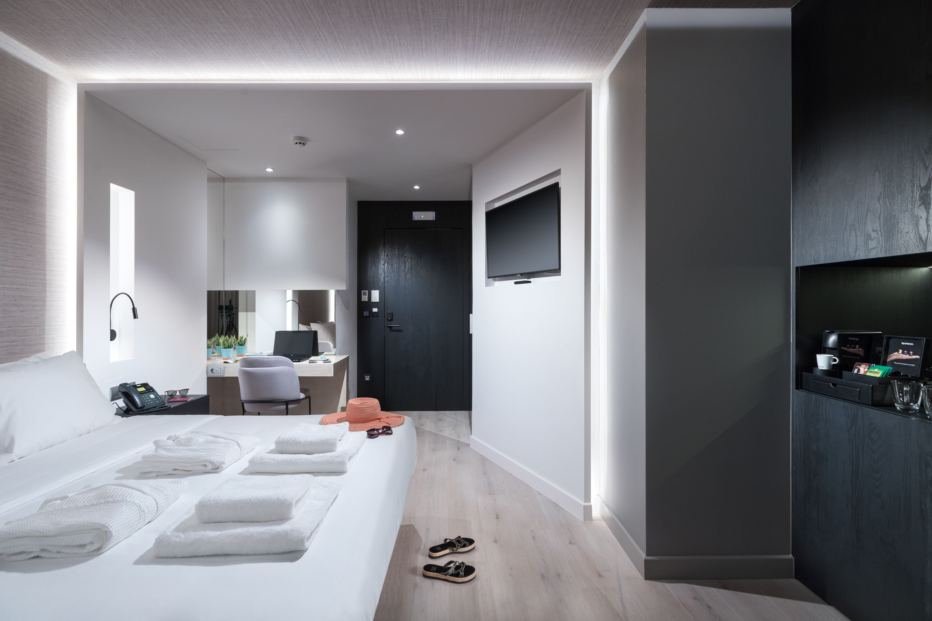 A modern hotel room with a neatly made bed, a desk with a computer, a TV mounted on the wall, and a coffee machine on the side table.