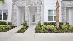 A modern two-story building with a stone facade, a central entrance, and a well-maintained garden with shrubs and a palm tree. - High luxury NEW 3 bedroom+loft townhouse in Hidden Forest, with private pool! (Clermont)