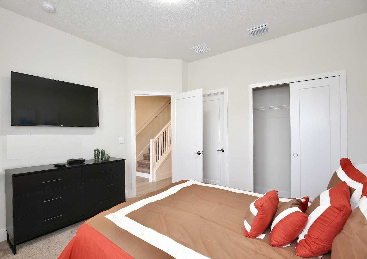 A bedroom with a bed, a TV mounted on the wall, a black dresser, and a staircase leading to another room.