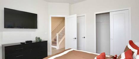 A bedroom with a bed, a TV mounted on the wall, a black dresser, and a staircase leading to another room.