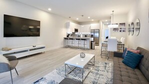 A modern living room with a flat-screen TV, a sofa, a coffee table, and a kitchen area in the background. - High luxury NEW 3 bedroom+loft townhouse in Hidden Forest, with private pool! (Clermont)