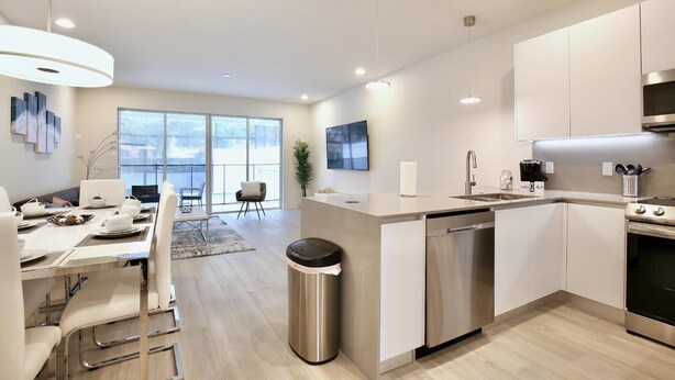A modern kitchen with a dishwasher, stainless steel appliances, and a dining area with a large window. - High luxury NEW 3 bedroom+loft townhouse in Hidden Forest, with private pool! (Clermont)