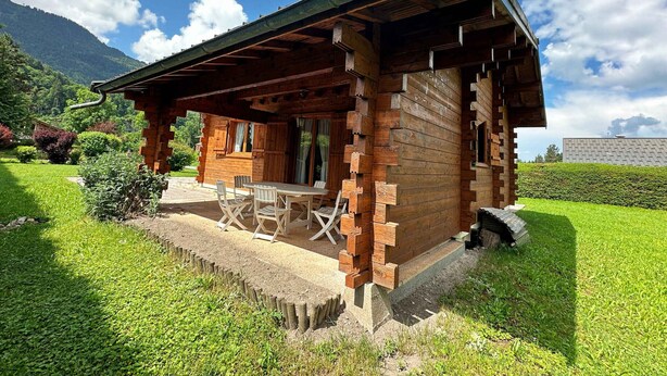 Eine Holzhütte mit Veranda und Essbereich, umgeben von üppiger Vegetation und Bergen.
