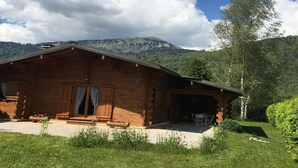 Eine Holzhütte mit Veranda, umgeben von üppiger Vegetation und Bergen im Hintergrund.