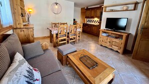 A cozy living room with a wooden sofa, a dining table with chairs, a wooden coffee table, a TV, and a kitchen area with wooden cabinets and a shelf.