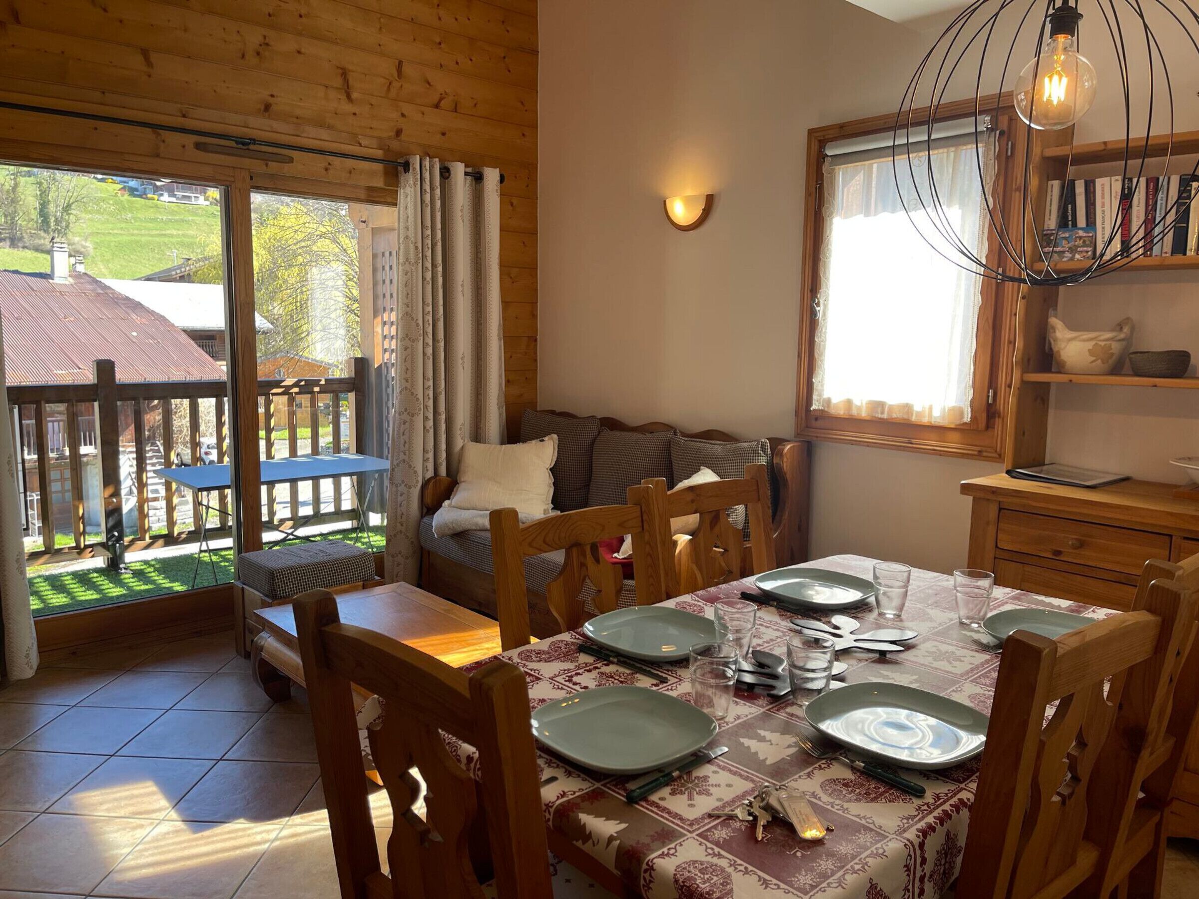 A cozy dining area with a wooden table set for four, a sofa, and a view of a garden through large windows.