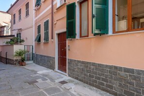 A narrow street with a building featuring a red door, green shutters, and potted plants.