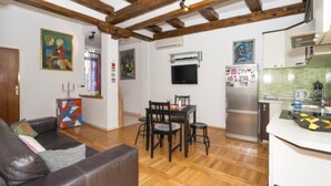 Un soggiorno con un divano in pelle marrone, un tavolo da pranzo in legno con sedie, una zona cucina con frigorifero e parete rivestita in piastrelle, e travi in legno al soffitto.