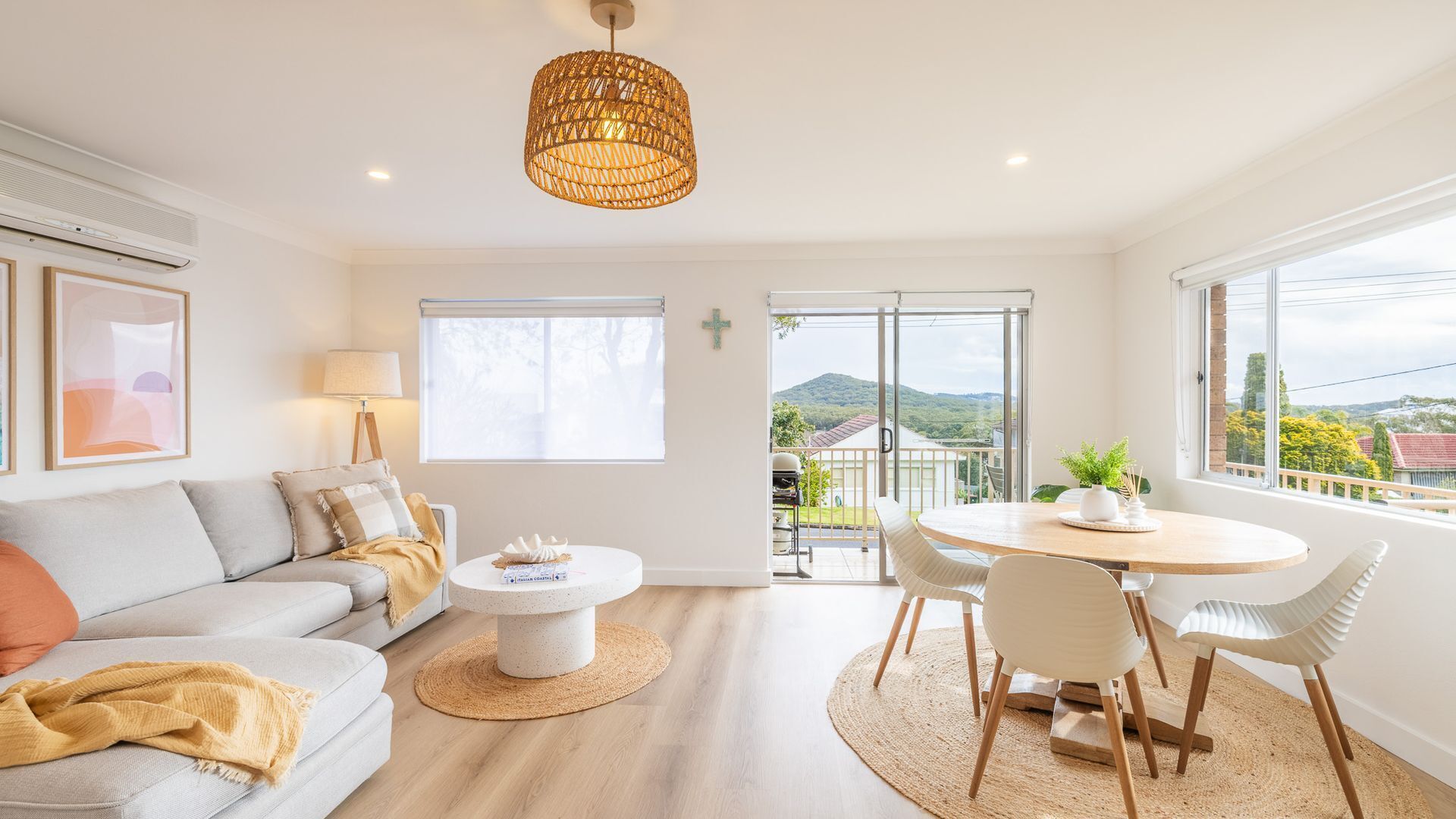 A modern living room with a sofa, coffee table, and dining area, featuring large windows and a view of the outdoors.