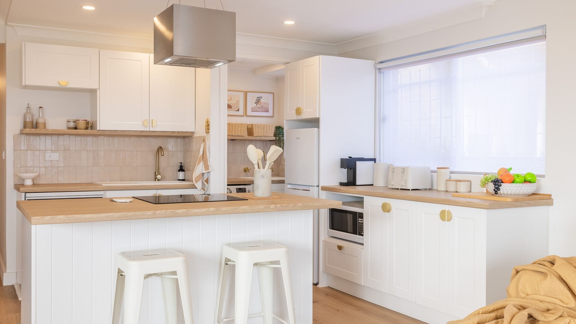 Una cocina moderna con armarios blancos, una isla central con taburetes, horno empotrado y un amplio ventanal que aporta luz natural.