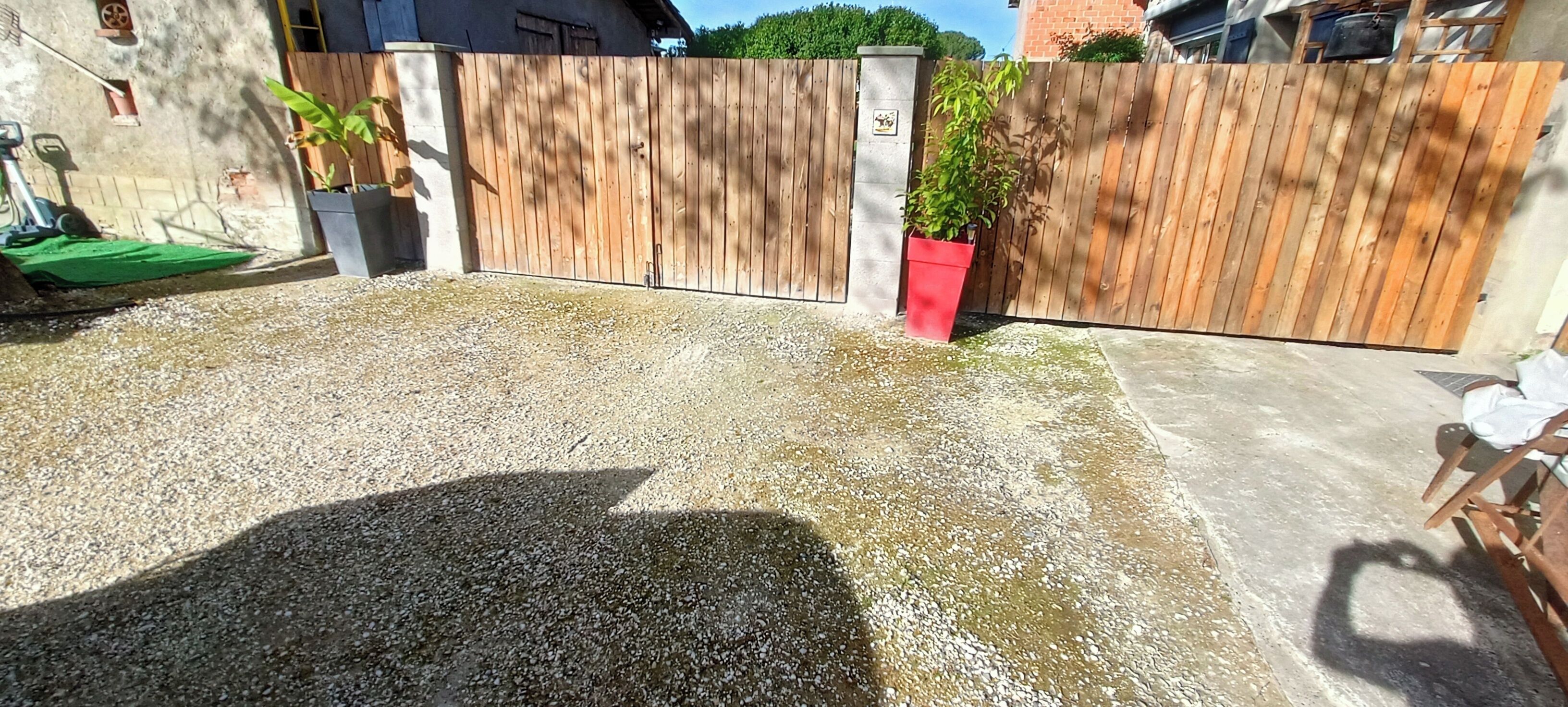 A gravel driveway leading to a wooden gate, with a red planter and a green mat near the gate.