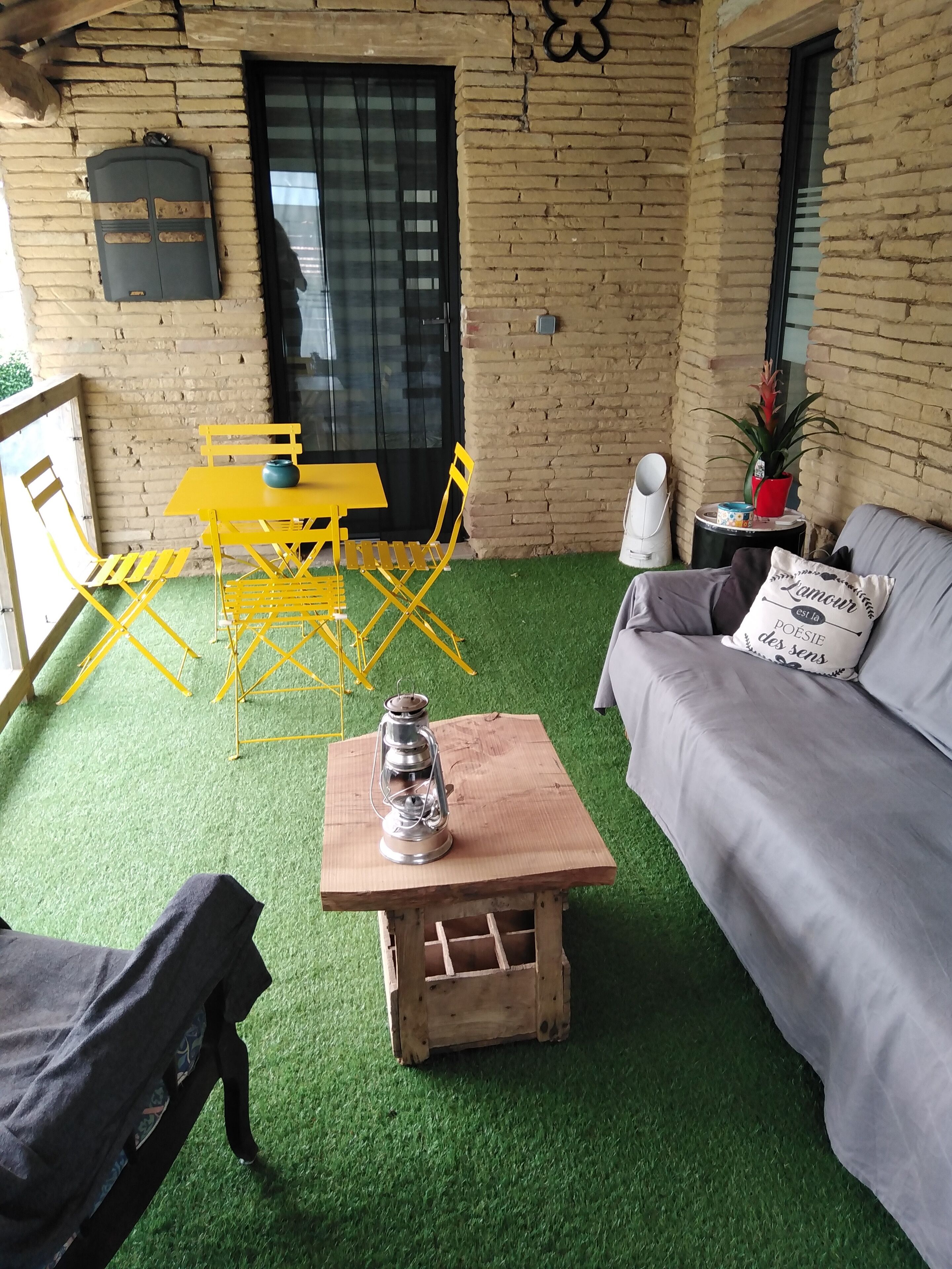 A cozy room with a green artificial turf floor, a wooden table with a lantern, a grey sofa with decorative pillows, and yellow folding chairs.