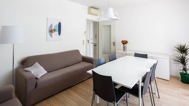 A modern living room with a brown sofa, a white dining table with chairs, a floor lamp, a wall art piece, and a potted plant.