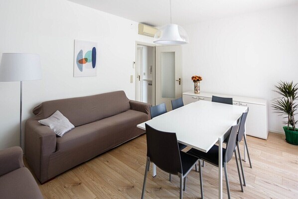 Un salon moderne avec un canapé marron, une table à manger blanche avec des chaises, un lampadaire, une œuvre d’art murale et une plante en pot.
