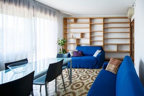 A living room with a blue sofa, a glass dining table, chairs, a patterned rug, and a wooden bookshelf.