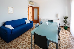 Ein Zimmer mit einem blauen Sofa, einem Esstisch mit Glasplatte und Stühlen, einer Kübelpflanze und gemusterten Bodenfliesen.