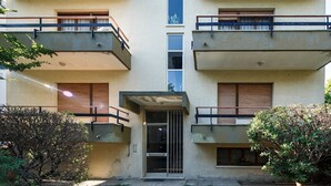 A multi-story building with balconies, a central entrance, and surrounding greenery.