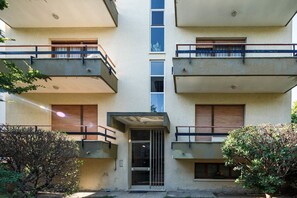 A multi-story building with balconies, a central entrance, and surrounding greenery.