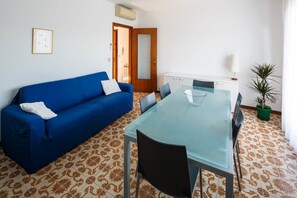 A room with a blue sofa, a glass-top dining table with chairs, a potted plant, and patterned floor tiles.